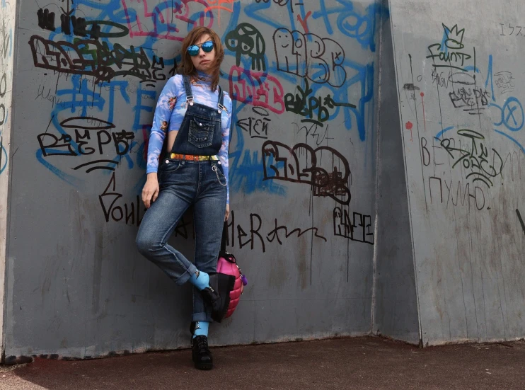 a woman leaning against a wall with graffiti on it, inspired by Max Gubler, tumblr, blue overalls, wearing blue sunglasses, neon steampunk, jean and multicolor shoes