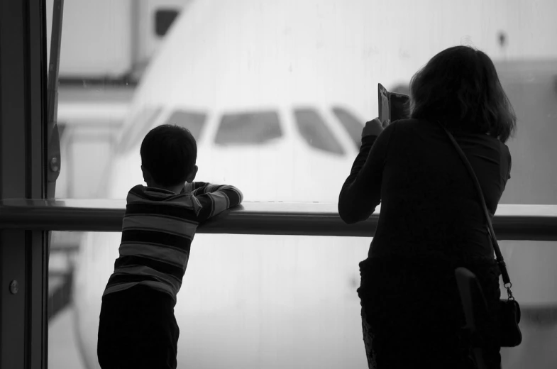a woman and a child looking out a window at an airplane, inspired by Elliott Erwitt, posing!!, telephoto shot, onlookers, waiting to strike