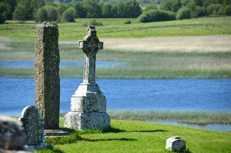 a couple of stone crosses sitting on top of a lush green field, a photo, by Edward Corbett, marshes, ancient tombs in the background, shoreline, looking across the shoulder