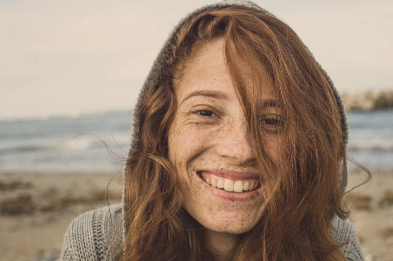 a close up of a woman with freckles on her face, a picture, with a beautifull smile, windy hair, at a beach, girl wearing hoodie