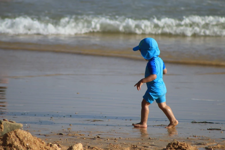 a little boy standing on top of a sandy beach, by Alison Debenham, pixabay, plasticien, blue outfit, side view of her taking steps, swimming to surface, wearing wide sunhat