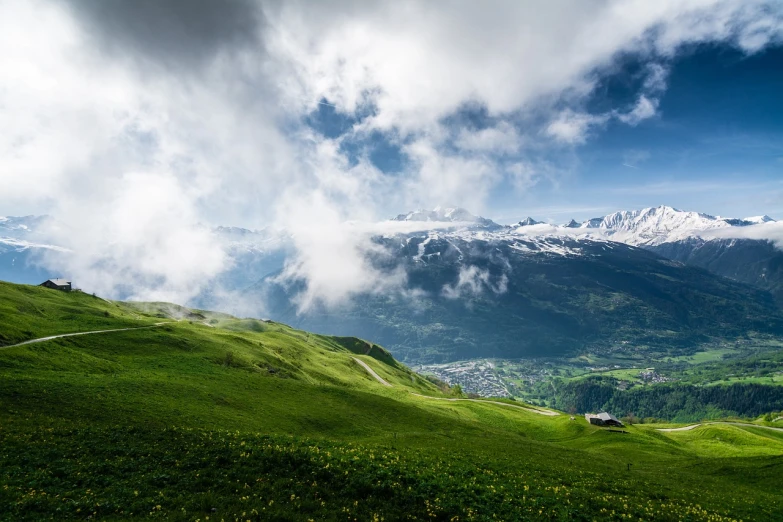 a view of the mountains from the top of a hill, by Cedric Peyravernay, nature and clouds in background, springtime morning, swiss alps, grass field surrounding the city