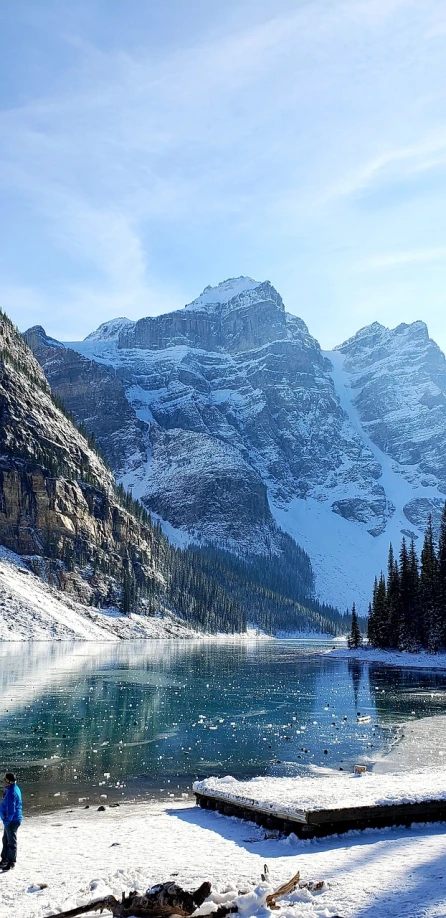 a man standing on top of a snow covered slope next to a lake, inspired by James Pittendrigh MacGillivray, pexels, giant imposing mountain, banff national park, closeup photo, wikimedia commons