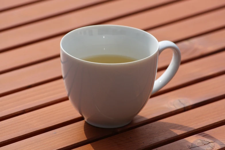 a cup of tea sitting on a wooden table, inspired by Wlodzimierz Tetmajer, pixabay, bauhaus, moringa juice, white mug, 7 0 mm photo, slightly sunny