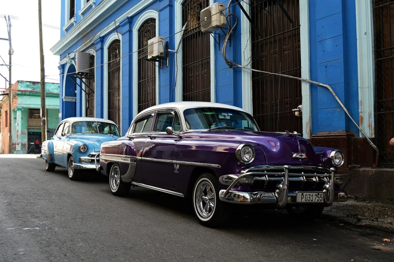 a row of classic cars parked in front of a blue building, by Juan Carlos Stekelman, flickr, dramatic purple thunders, cuban revolution, two - tone, standing in front of lowrider