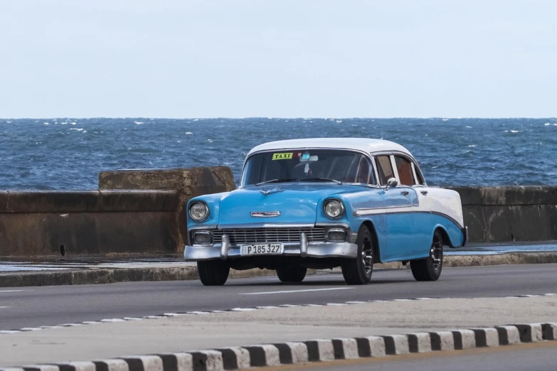 a blue and white car driving down a road next to the ocean, a portrait, by Tom Wänerstrand, flickr, che guevara, 5 hotrods driving down a street, 8k h- 640, taxi