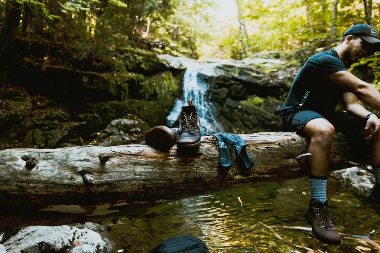 a man sitting on a log in front of a waterfall, by Alexander Brook, unsplash, riding boots, william penn state forest, clothes floating, 33mm photo