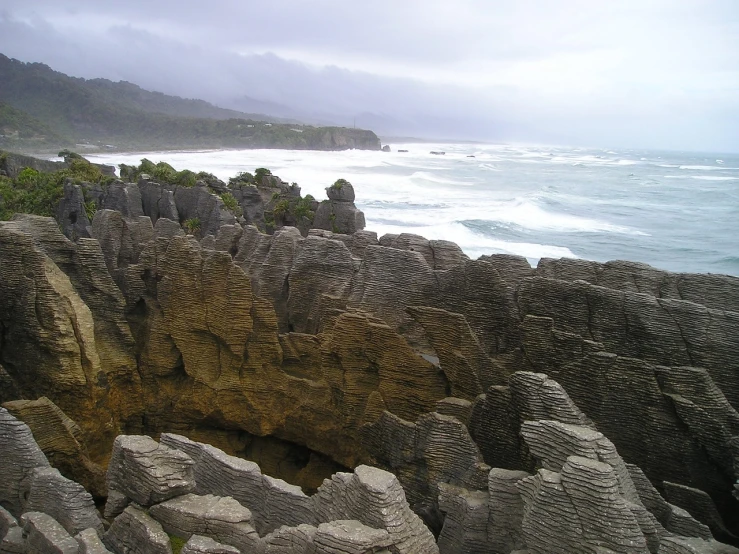 a man standing on top of a rocky cliff next to the ocean, by Alexander Scott, flickr, hurufiyya, overcast!!!, chiseled formations, kahikatea, panels