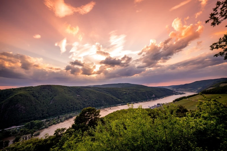 a river flowing through a lush green valley under a cloudy sky, a stock photo, by Matthias Weischer, shutterstock, romanticism, pink sunset, germany. wide shot, dramatic reddish light, looking over west virginia