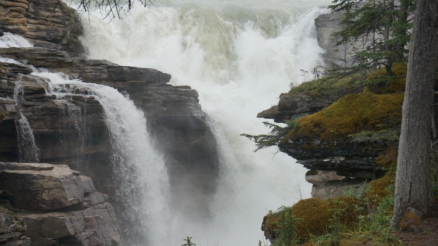 a man standing on top of a cliff next to a waterfall, rushing water, photo taken from far away, bc, 8 feet fall