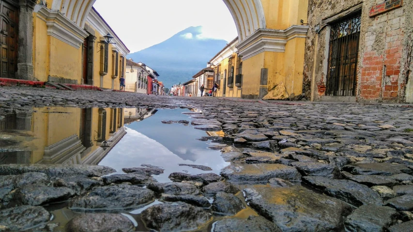 a puddle of water on a cobblestone street, a picture, by Andrée Ruellan, pexels, quito school, volcano setting, arch, view from below, 🪔 🎨;🌞🌄