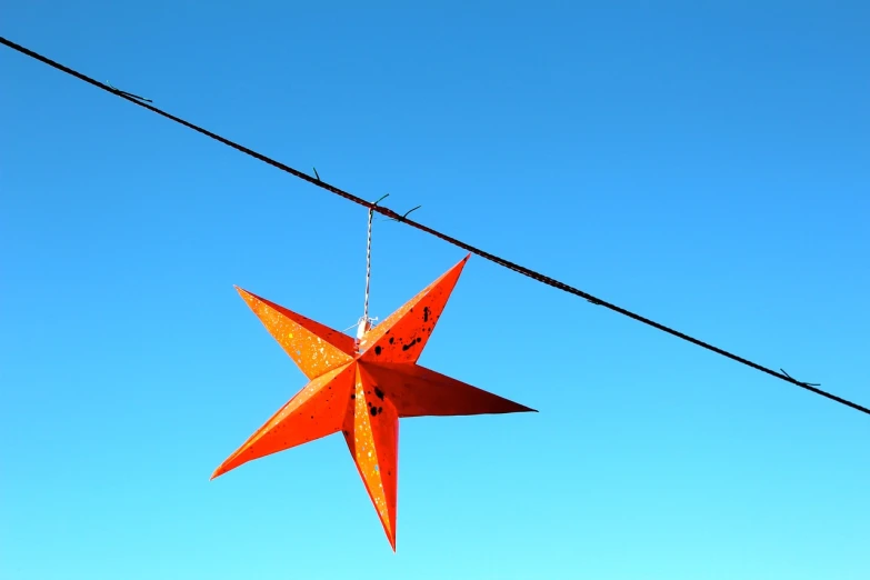 a star hanging from a wire with a blue sky in the background, a photo, by Erwin Bowien, flickr, folk art, orange lamp, sharp lighting. bright color, red and orange colored, holiday