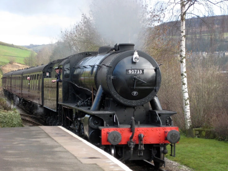 a train traveling down train tracks next to a lush green hillside, a portrait, by Dave Allsop, brass and steam technology, black mountains, winter sun, round-cropped