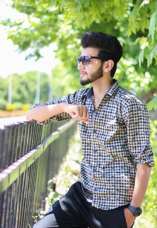 a man in a checkered shirt leaning on a fence, a picture, inspired by Jitish Kallat, designer sunglasses, islamic, beautiful sunny day, full - view