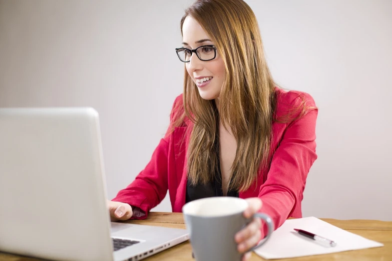 a woman sitting at a table with a laptop and a cup of coffee, girl wearing round glasses, well lit professional photo, welcoming grin, in front of a computer