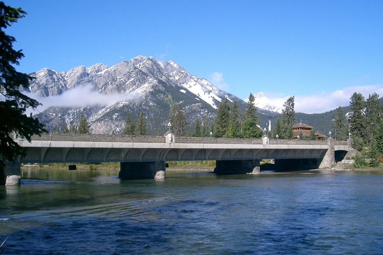 a bridge over a river with mountains in the background, by Brigette Barrager, flickr, big sky, wikipedia, british columbia, concrete