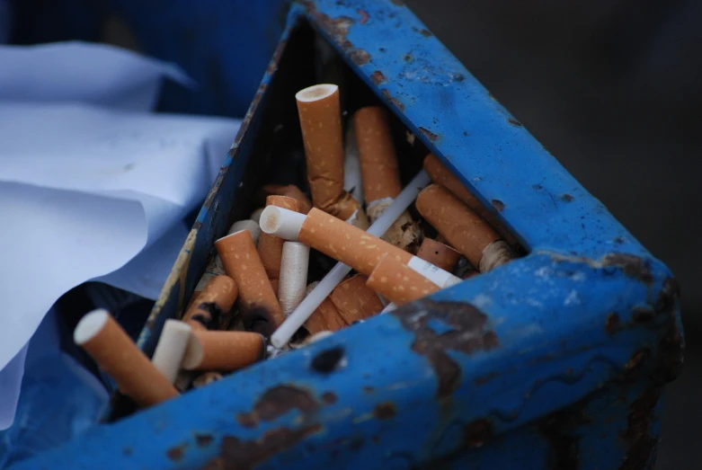 a blue trash can filled with lots of cigarettes, smoking vessels, up close picture, hard lines, holding a cigarette