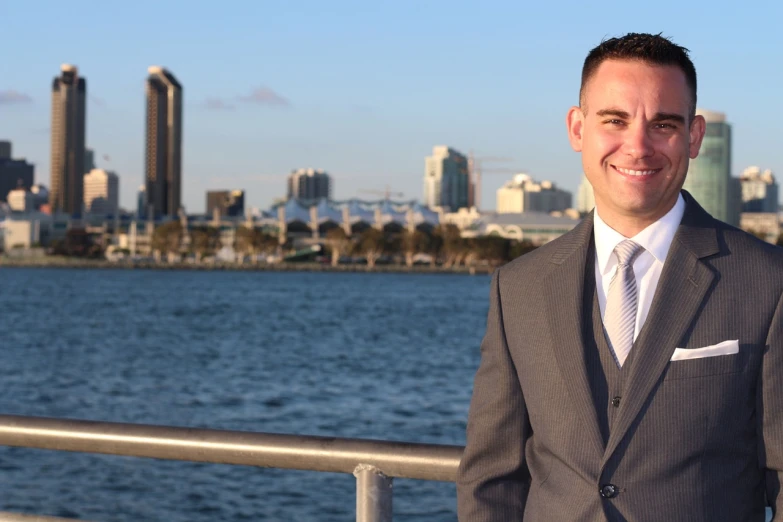 a man in a suit standing in front of a body of water, a portrait, by Robbie Trevino, flickr, long beach background, city skyline in the background, taking control while smiling, video still