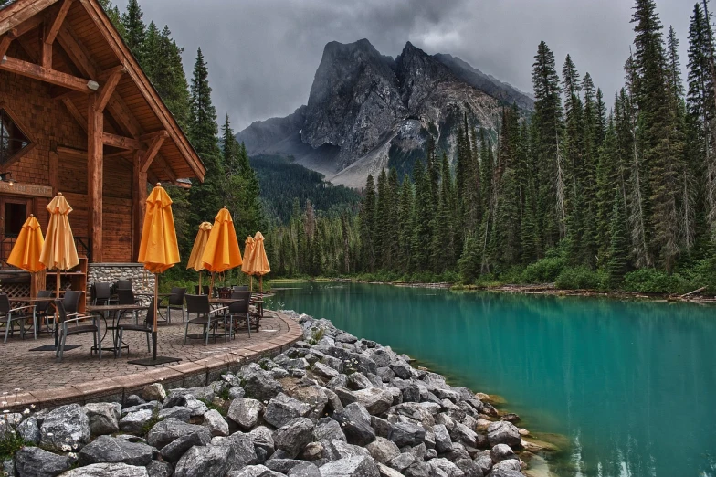 a cabin with tables and umbrellas next to a body of water, by Erik Pevernagie, pexels contest winner, banff national park, [[fantasy]], lush vista, rocky mountains