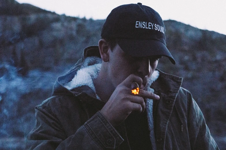 a man smoking a cigarette in front of a mountain, by Brian Snøddy, unsplash, digital art, faded hat, lowkey lighting, streetwear, 35 mm product photo”