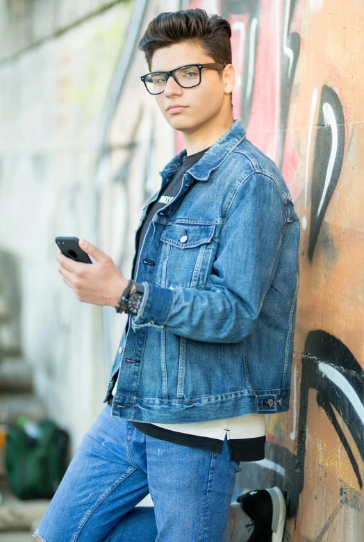 a man leaning against a wall with a cell phone, a picture, inspired by Jean Malouel, pexels, realism, denim jacket, jewish young man with glasses, high quality details, high resolution