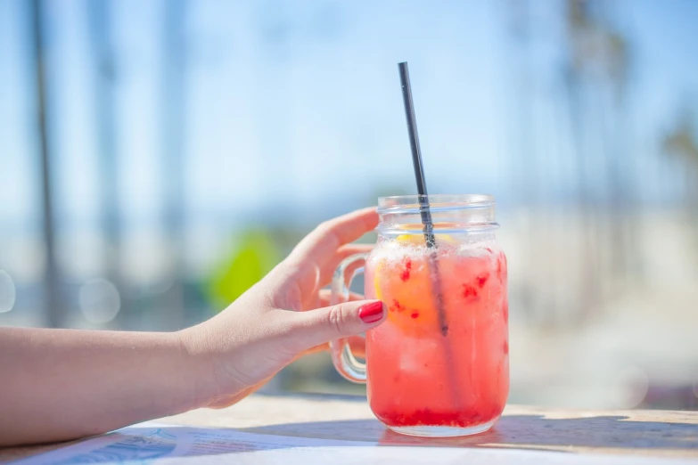 a person holding a glass with a drink in it, a stock photo, sunny day at beach, pink and red color style, back of hand on the table, jackstraws