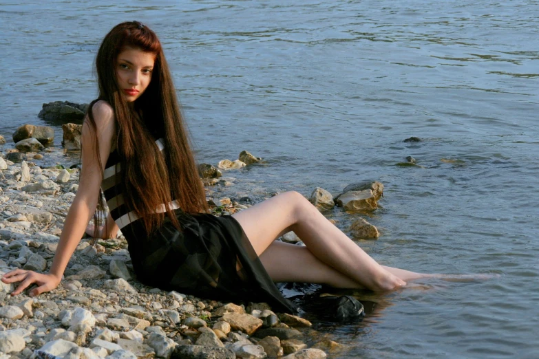 a woman sitting on a rocky beach next to a body of water, a portrait, inspired by Ksenia Milicevic, flickr, renaissance, 1 7 - year - old goth girl, full body:: sunny weather::, loosely cropped, summer evening