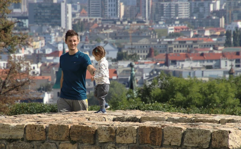 a man and a little girl standing on top of a stone wall, by Adam Szentpétery, happening, city in backround, 30 year old man, toddler, not cropped