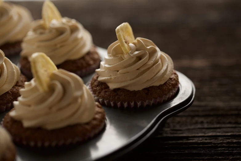 a white plate topped with cupcakes covered in frosting, by Etienne Delessert, banana, light brown, on a dark background, on a wooden tray