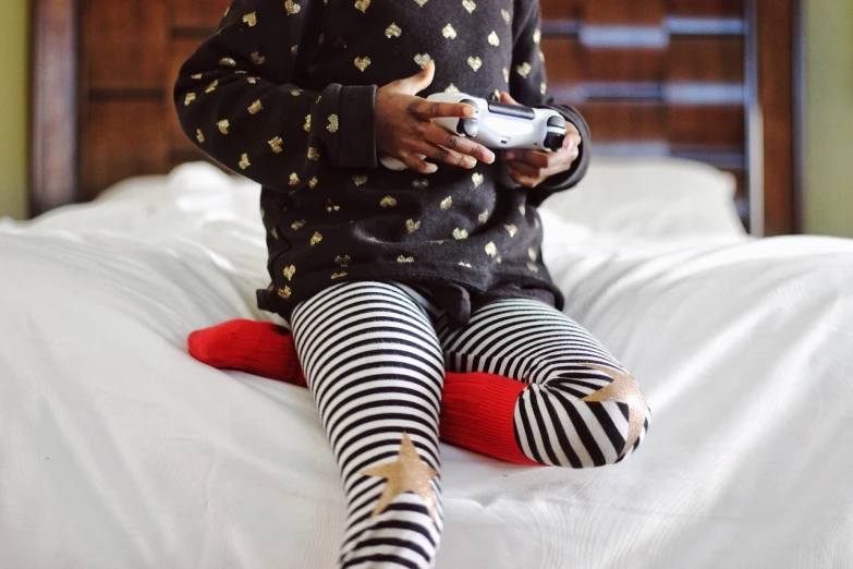 a little girl sitting on top of a bed holding a video game controller, by David Garner, pexels, striped pantyhose, red gold and black outfit, black skin, polka dot