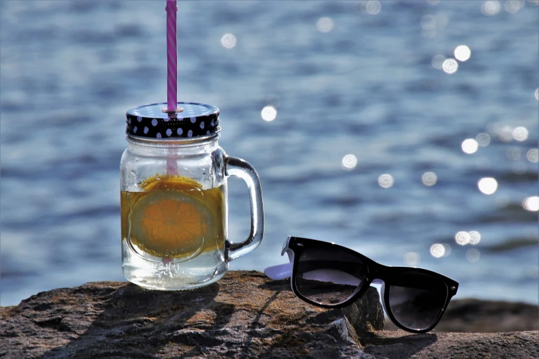 a drink in a mason jar next to a pair of sunglasses, a picture, by Mathias Kollros, pexels, at the waterside, “ golden cup, black rimmed glasses, lemonlight