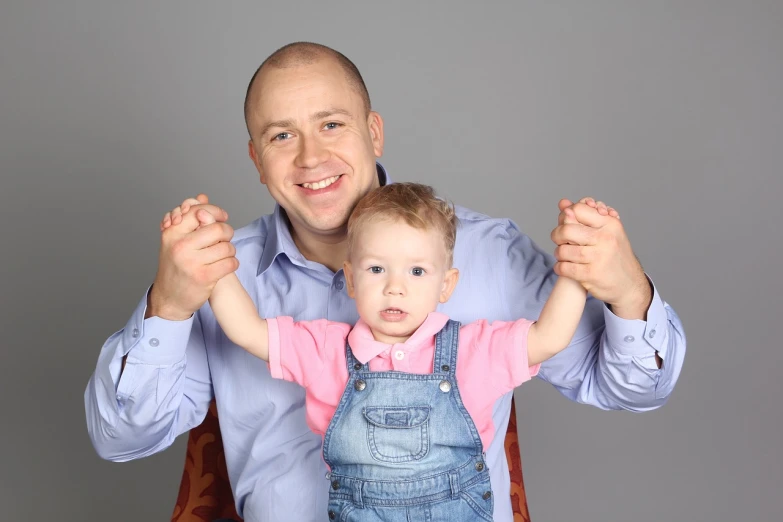 a man holding a baby in his arms, a portrait, shutterstock, studio portrait photo, waving at the camera, alex maksiov and john pugh, bald on top