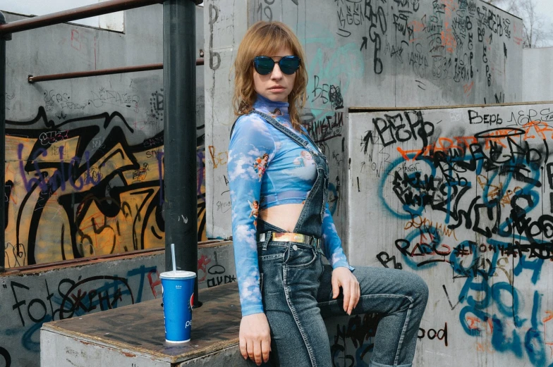 a woman standing in front of a graffiti covered wall, inspired by Elsa Bleda, graffiti, wearing blue jean overalls, imogen poots, wearing versace sunglasses, “gas station photography