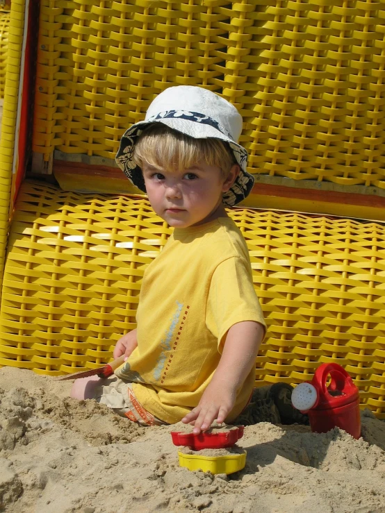 a little boy that is sitting in the sand, by Dietmar Damerau, flickr, yellow hardhat, small blond goatee, softplay, red!! sand
