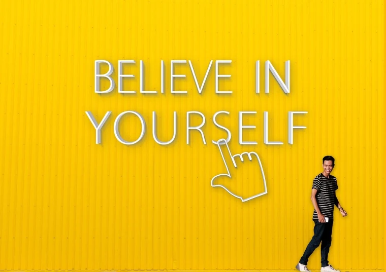 a man standing in front of a yellow wall that says believe in yourself, a picture, inspired by Ryan Yee, pexels, thumb up, wax figure, billboard image, high resolution print :1 cmyk :1