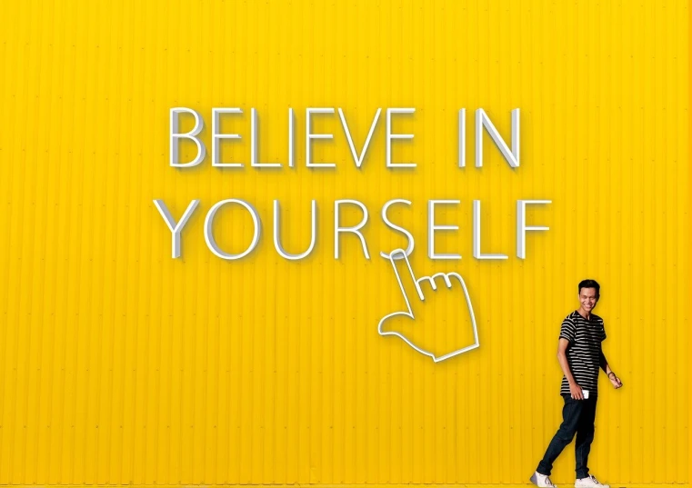 a man standing in front of a yellow wall that says believe in yourself, a picture, inspired by Ryan Yee, pexels, thumb up, wax figure, billboard image, high resolution print :1 cmyk :1