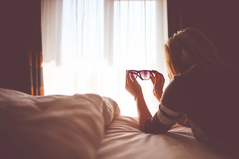 a woman sitting on top of a bed holding a pair of sunglasses, a picture, by Matija Jama, pexels, istock, back - shot, pink glasses, early in the morning