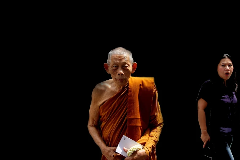 a man in an orange robe standing next to a woman, by Richard Carline, minimalism, thai, wise old man, on black background, reuters