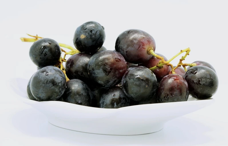 a white bowl filled with black grapes on top of a table, pixabay, set against a white background, background image, drops, stacked image