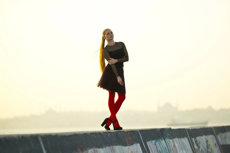 a woman standing on top of a cement wall, a picture, inspired by Elsa Bleda, shutterstock, renaissance, in red velvet stockings, long yellow hair, full body backlight, on a bridge