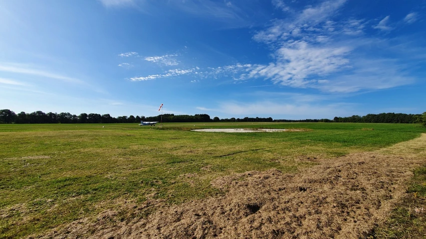 a man flying a kite on top of a lush green field, a picture, by Robert Zünd, land art, lots of mud puddles and craters, on a landing pad, clear blue skies, shot with iphone 1 0