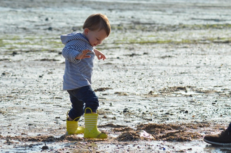a little boy standing on top of a muddy field, yellow seaweed, wearing boots, walking on the beach, milk puddles