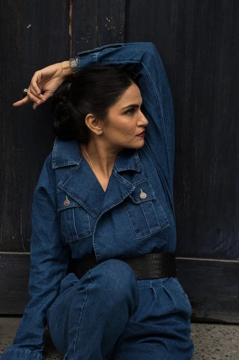 a woman in a denim jumpsuit sitting on the ground, style of ade santora, shohreh aghdashloo, hands behind her pose!, leaning on the wall