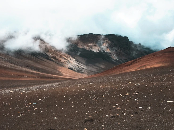 a person standing in the middle of a desert with a mountain in the background, a tilt shift photo, by Anna Haifisch, trending on unsplash, color field, maui, flora-lush-crater, covered in clouds, gravel and scree ground