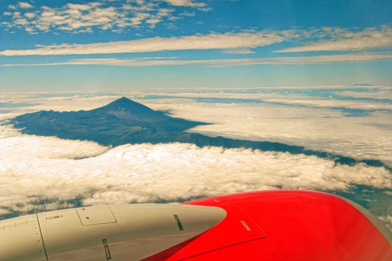 an airplane flying above the clouds with a mountain in the background, a photo, by Juan O'Gorman, flickr, precisionism, lava!!!, red wings, volcanos, viewed from bird's-eye