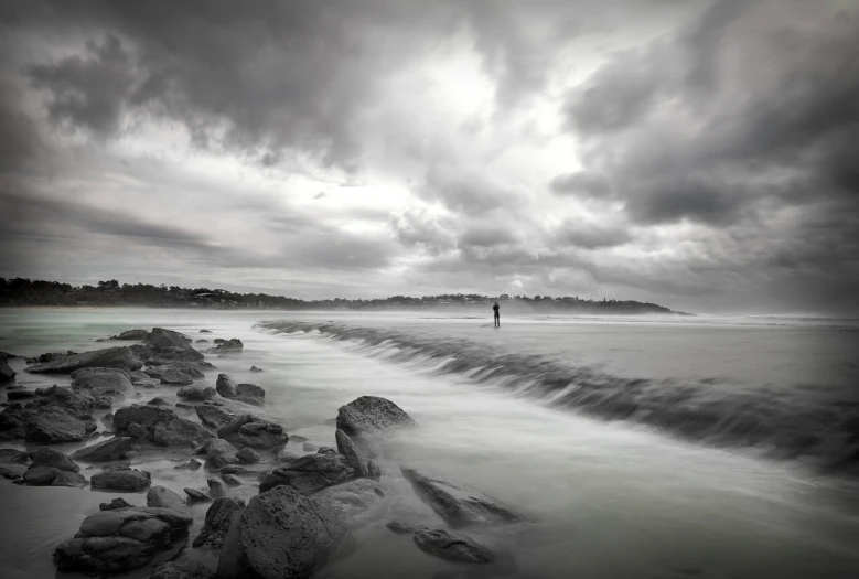 a man standing on top of a rocky beach next to the ocean, a black and white photo, by Etienne Delessert, violent stormy waters, manly, time - lapse, overcast dusk