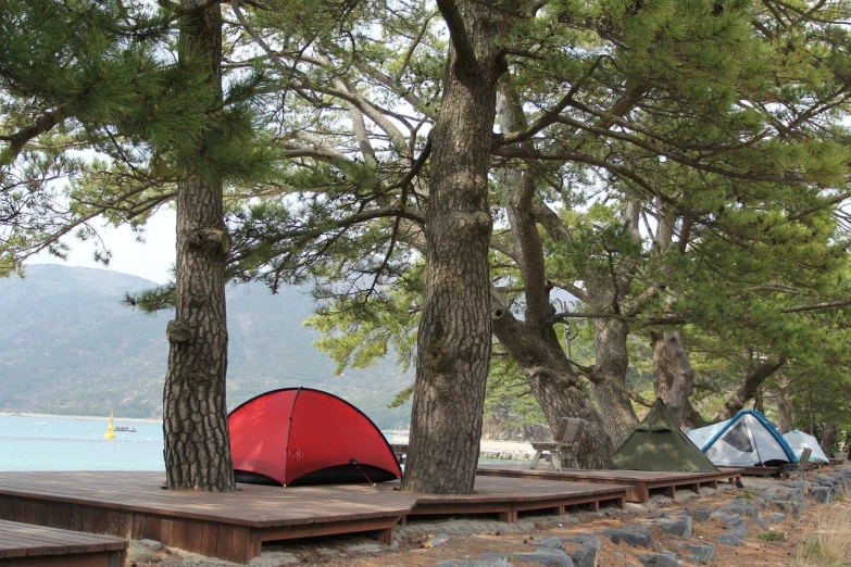 a red tent sitting on top of a wooden deck, by Kiyohara Tama, shutterstock, shin hanga, beach trees in the background, tent camp in foreground, greece, with dark trees in foreground