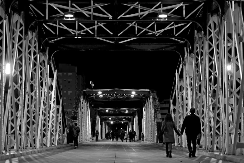 a group of people walking across a bridge at night, a black and white photo, by Andrei Kolkoutine, symmetric!!, in a massive cavernous iron city, ponte 2 5 de abril, archway