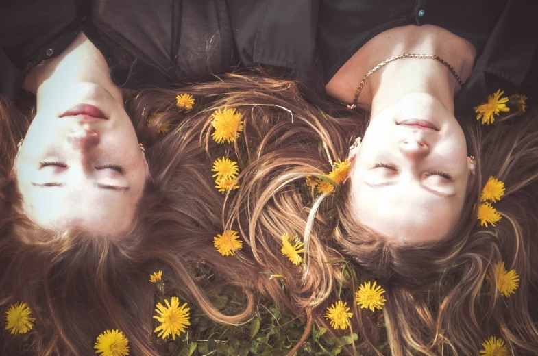 a couple of women laying on top of a lush green field of flowers, a picture, pexels, romanticism, two identical symmetrical eyes, dandelions, floating long hair, [ realistic photography ]