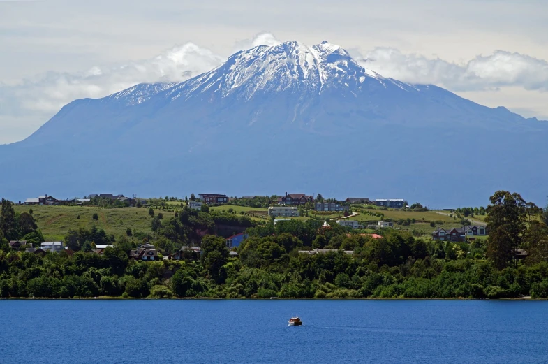 a boat in a body of water with a mountain in the background, flickr, hurufiyya, volcano in background, in the foreground a small town, tula, with snow on its peak